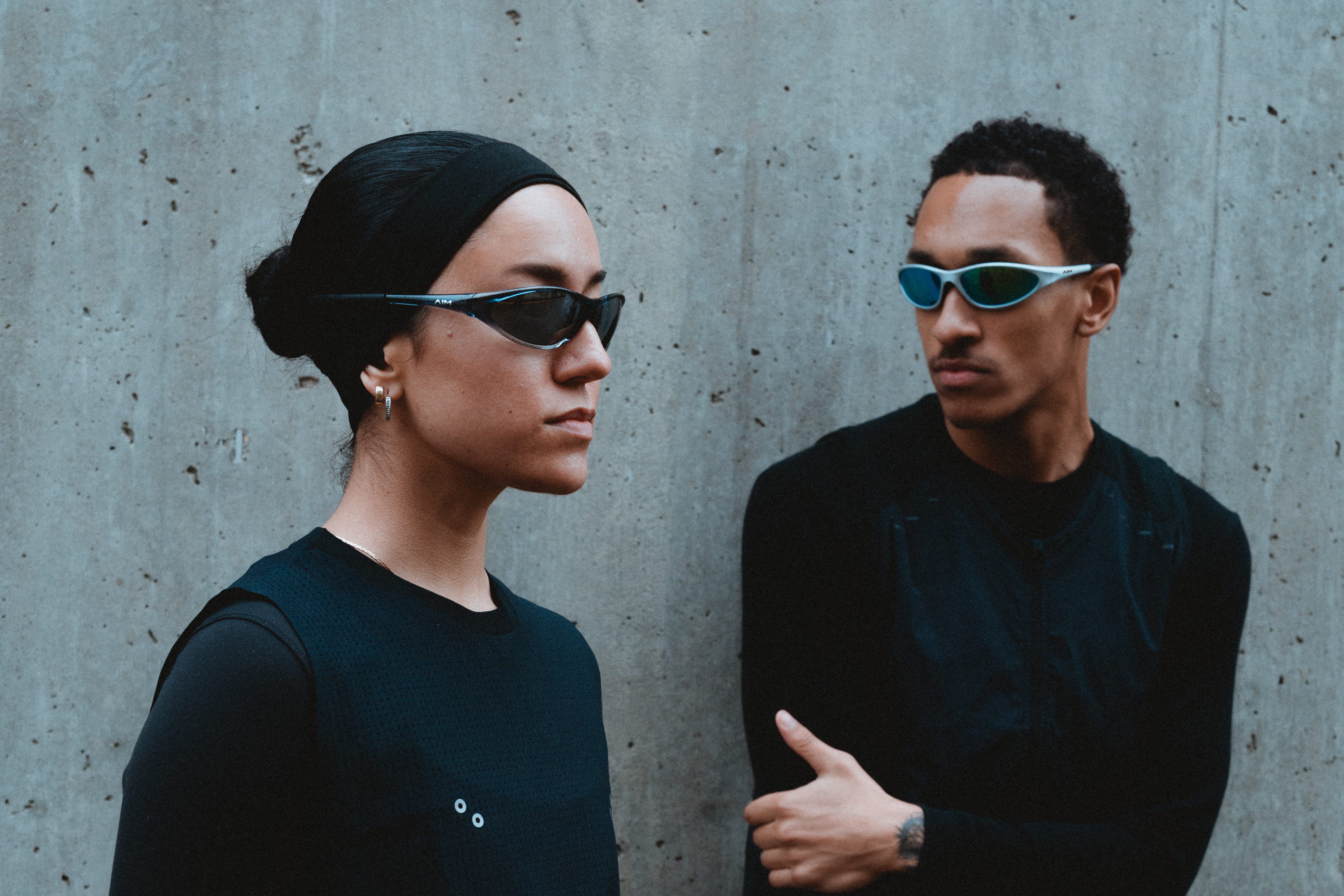 Two people wearing dark clothing and sunglasses standing against a concrete wall.